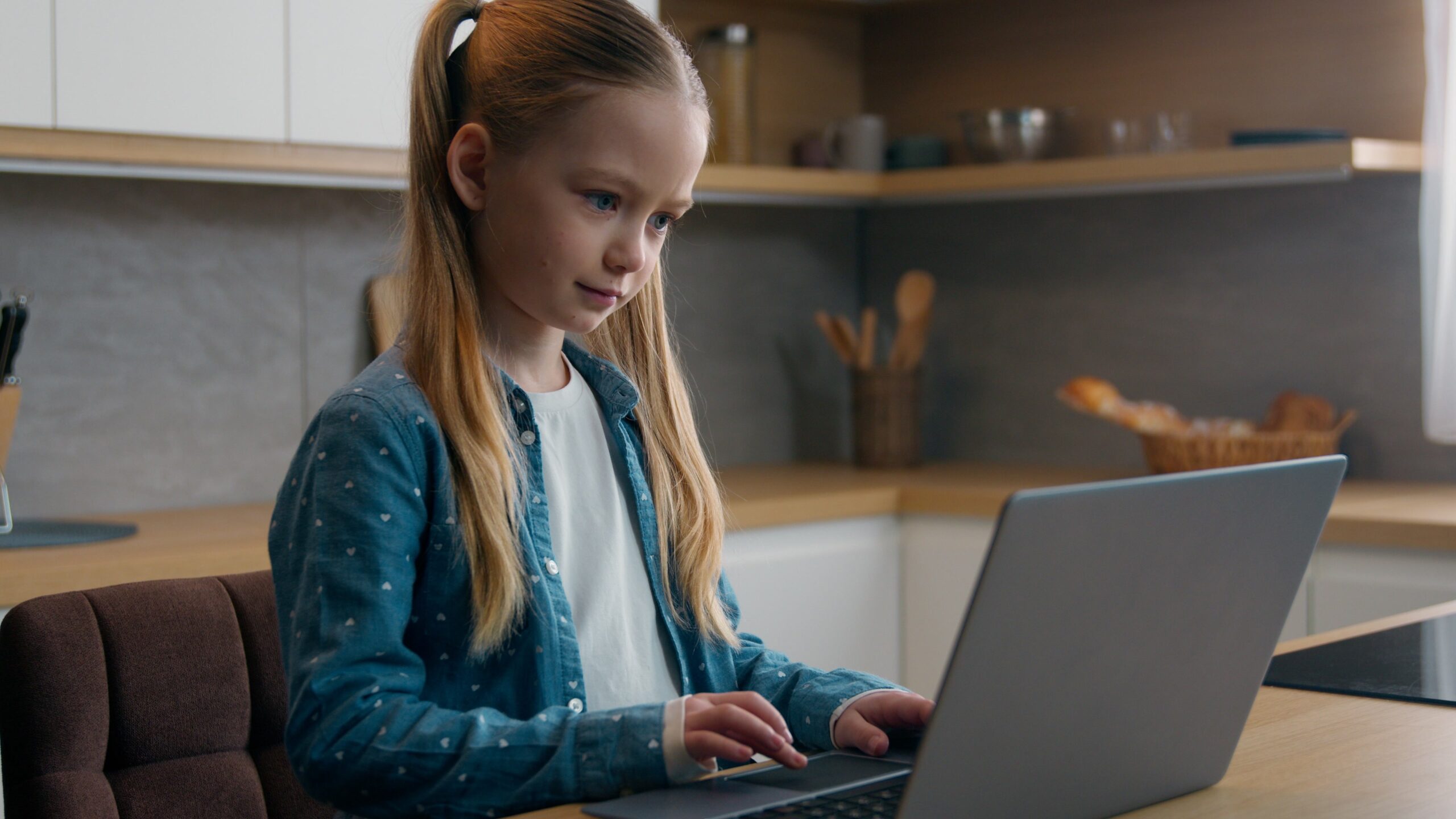 A young girl with long blonde hair in a ponytail sits at a kitchen table, focused on using a laptop. The kitchen background includes shelves, utensils, and neutral-colored decor.